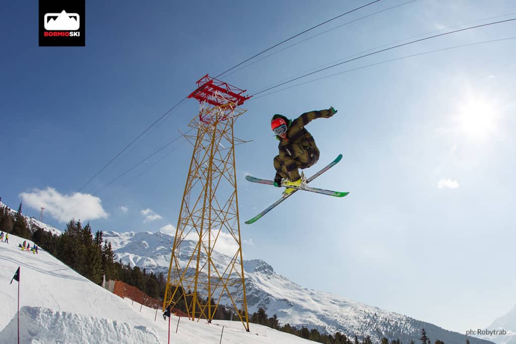 Snowpark Bormio aperto. Sciatore salta dal kicker con sci incrociati e presa grab.